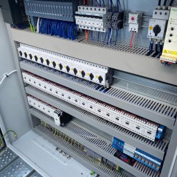 Cirqo Instalacje Elektryczne Bartłomiej Musiał - Otwarta szafa sterownicza z widocznymi modułami PLC Siemens, bezpiecznikami Eaton, stycznikami i okablowaniem w kolorach niebieskim, czerwonym i czarnym. Widoczna perforowana konstrukcja wsporcza...