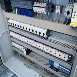 Otwarta szafa sterownicza z widocznymi modułami PLC Siemens, bezpiecznikami Eaton, stycznikami i okablowaniem w kolorach niebieskim, czerwonym i czarnym. Widoczna perforowana konstrukcja wsporcza...