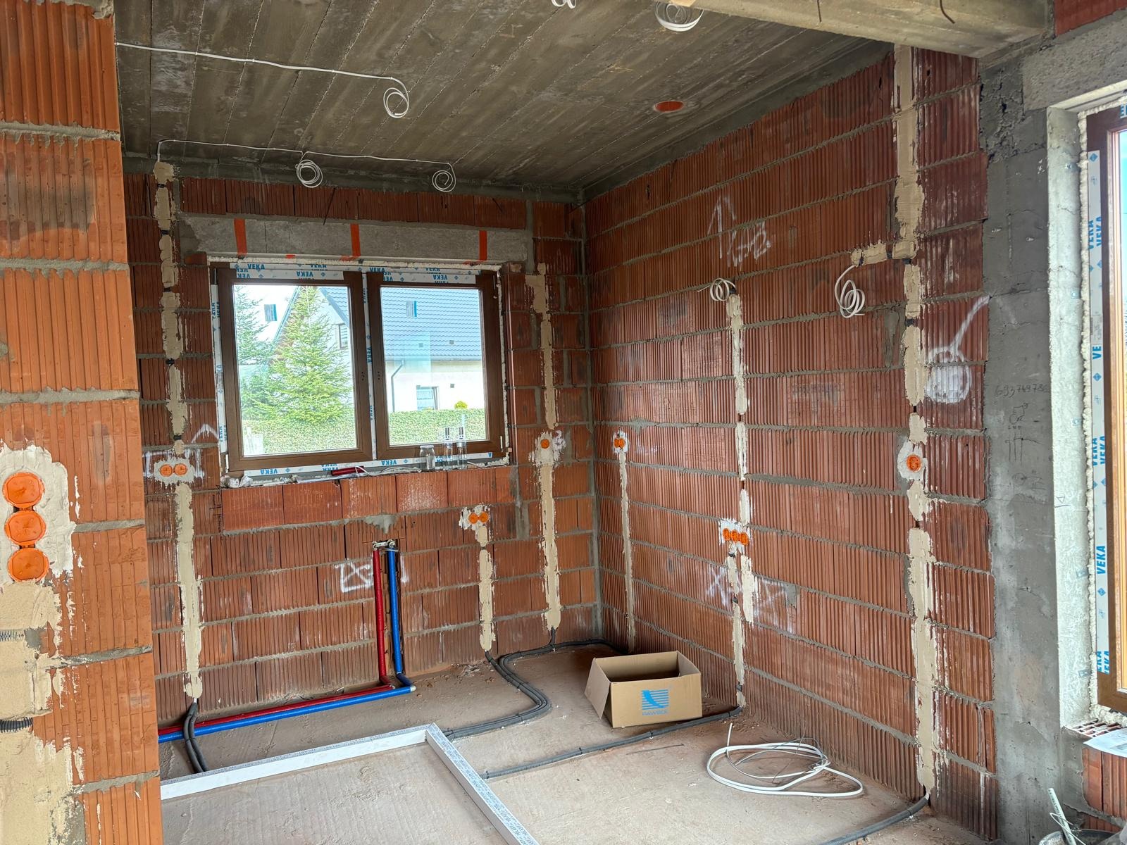 Wnętrze w trakcie prac instalacyjnych: widoczne przewody elektryczne w ścianach z cegły, puszki instalacyjne, rury wodne w kolorach niebieskim i czerwonym oraz częściowo surowy sufit betonowy.