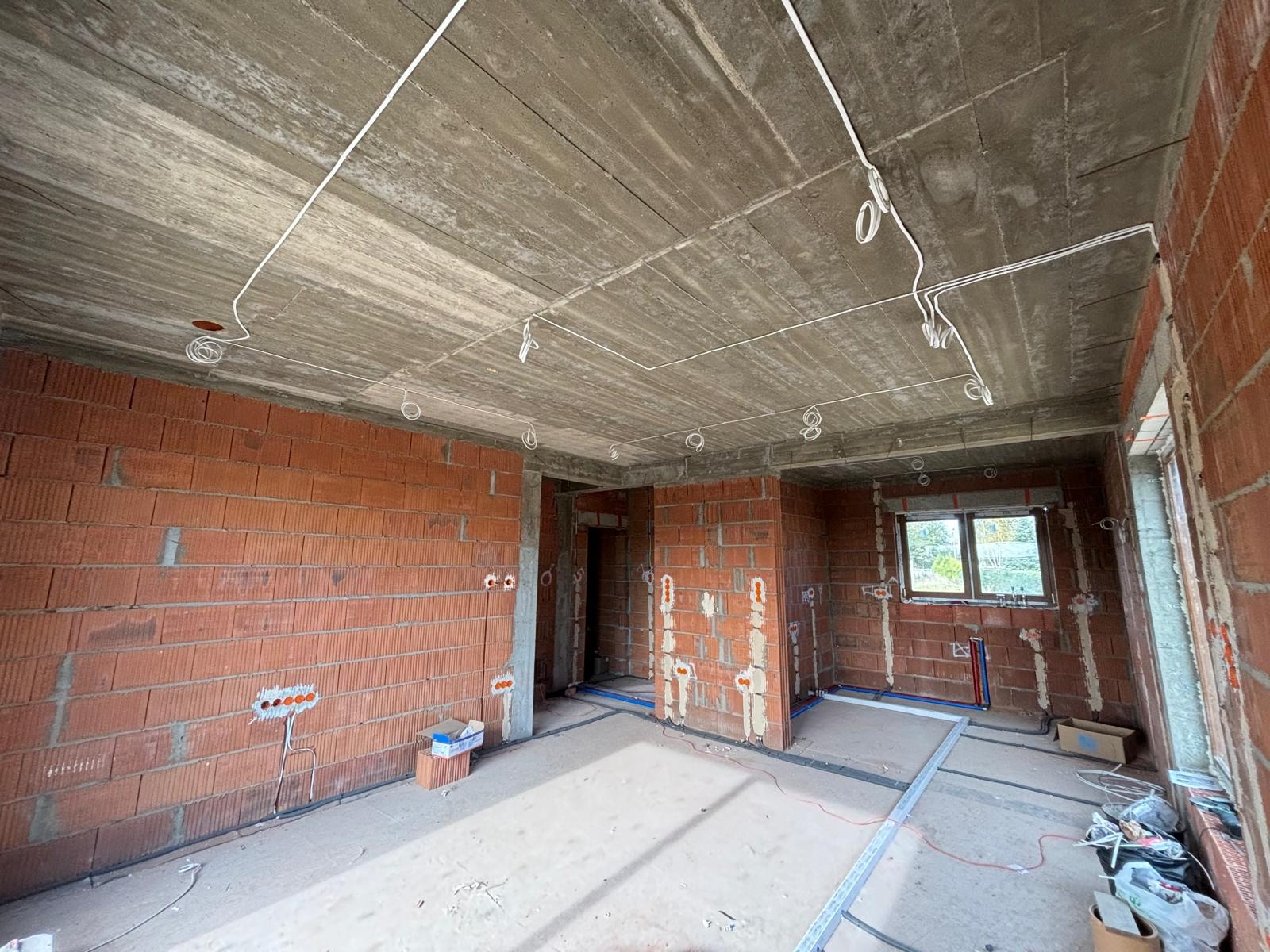 Surowy stan wnętrza budynku w trakcie prac elektrycznych: widoczne kable rozprowadzone po suficie, puszki elektryczne w ścianach z czerwonej cegły, oraz instalacja wodna z rurami w kolorze...