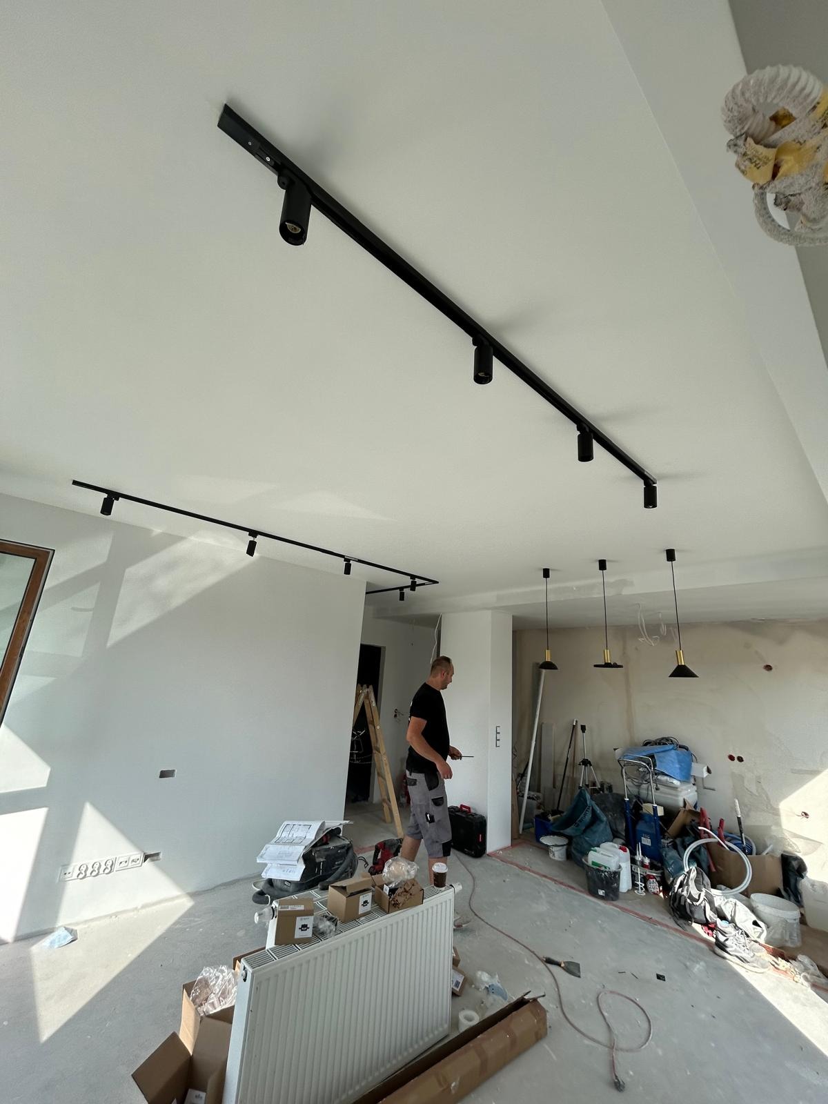 Montaż szynoprzewodów z reflektorami w nowym mieszkaniu, widoczny elektryk z narzędziami, surowy stan wykończenia wnętrza, okablowanie zwisające z sufitu w tle.