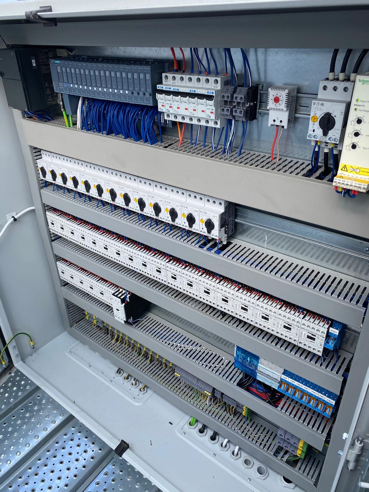 Otwarta szafa sterownicza z widocznymi modułami PLC Siemens, bezpiecznikami Eaton, stycznikami i okablowaniem w kolorach niebieskim, czerwonym i czarnym. Widoczna perforowana konstrukcja wsporcza...