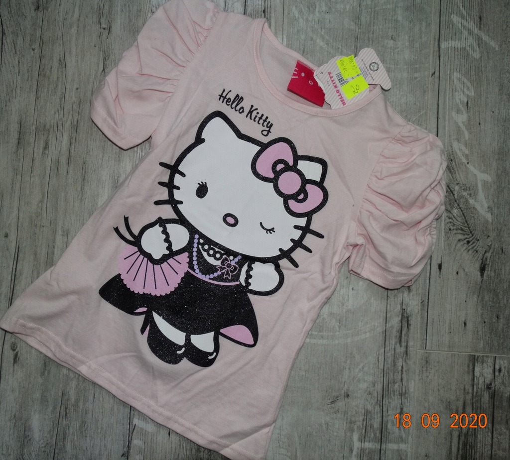 Jasnoróżowa koszulka dziewczęca z krótkimi, marszczonymi rękawami i nadrukiem Hello Kitty w czarnej, brokatowej sukience z wachlarzem, leżąca ukośnie na drewnianym tle.