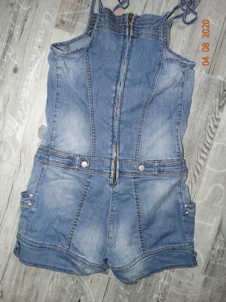 Używany dżinsowy kombinezon damski z rozpiętym zamkiem błyskawicznym na plecach, leżący na podłodze z paneli.