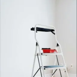 Biała drabina z czerwonym kuwetą i wałkiem malarskim na tle świeżo pomalowanej, jasnej ściany w rogu pomieszczenia, zabezpieczona podłoga.