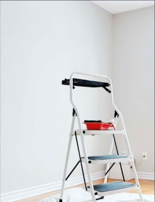 Biała drabina z czerwonym kuwetą i wałkiem malarskim na tle świeżo pomalowanej, jasnej ściany w rogu pomieszczenia, zabezpieczona podłoga.