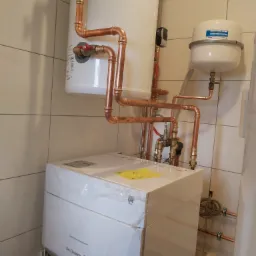 Nowoczesny, biały piec gazowy De Dietrich z podłączonymi miedzianymi rurami, zaworami i zbiornikiem wyrównawczym, zainstalowany w pomieszczeniu wyłożonym jasnymi płytkami.