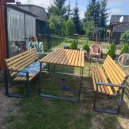 Zestaw mebli ogrodowych z drewnianymi blatami i siedziskami na metalowych, ciemnych ramach, ustawiony na trawniku w ogrodzie.