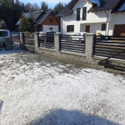 Nowoczesne ogrodzenie posesji z poziomymi przęsłami i słupkami z szarego kamienia, widoczne dwa budynki mieszkalne i zaparkowane samochody, podłoże pokryte śniegiem.