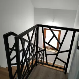 Czarna, geometryczna balustrada schodowa z metalu, widok z góry, drewniane schody i okno w tle.