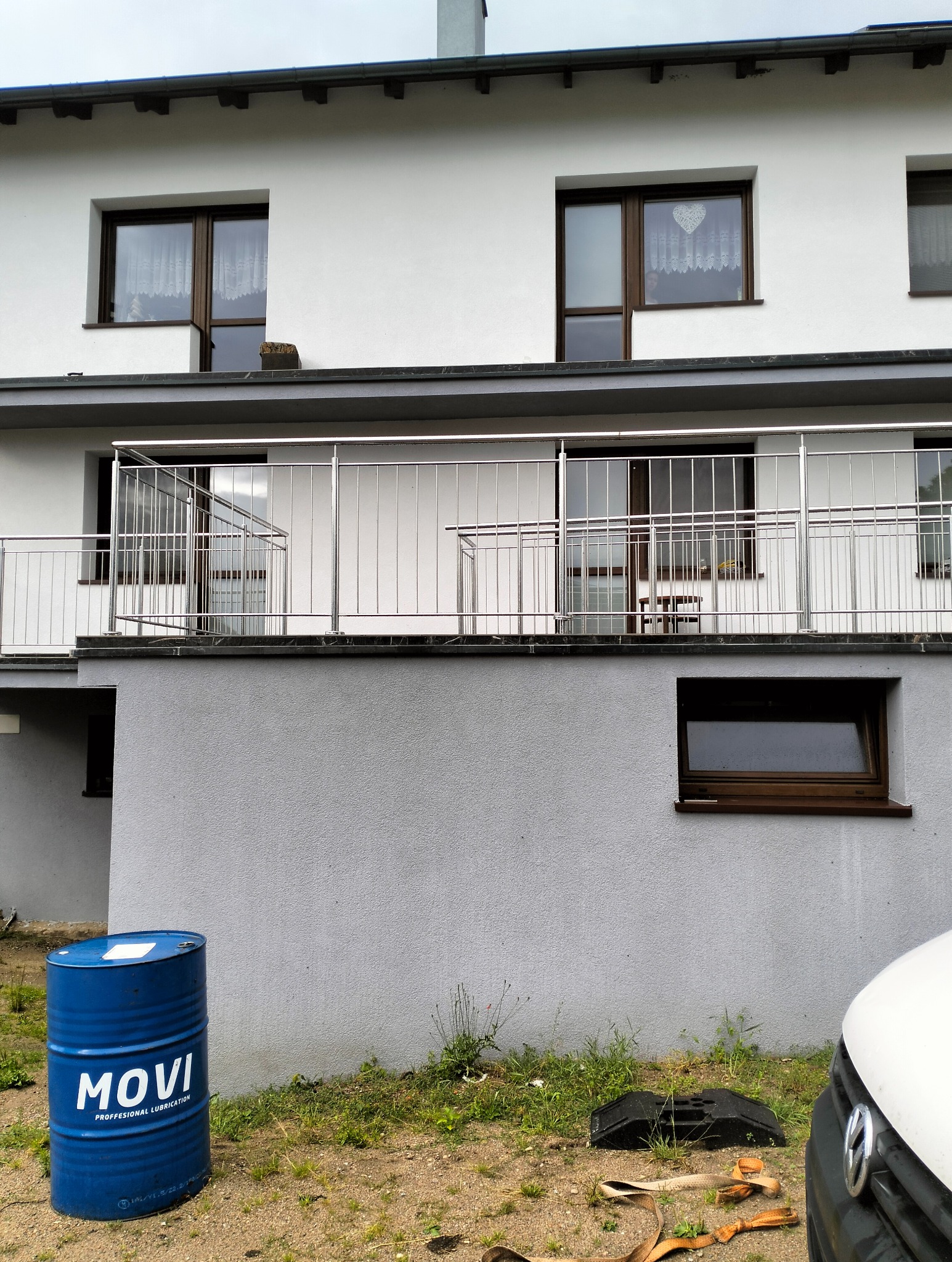 Dwupiętrowy budynek z balkonami zabezpieczonymi nowoczesnymi, nierdzewnymi balustradami o minimalistycznym designie, z widoczną beczką oleju i fragmentem samochodu na pierwszym planie.