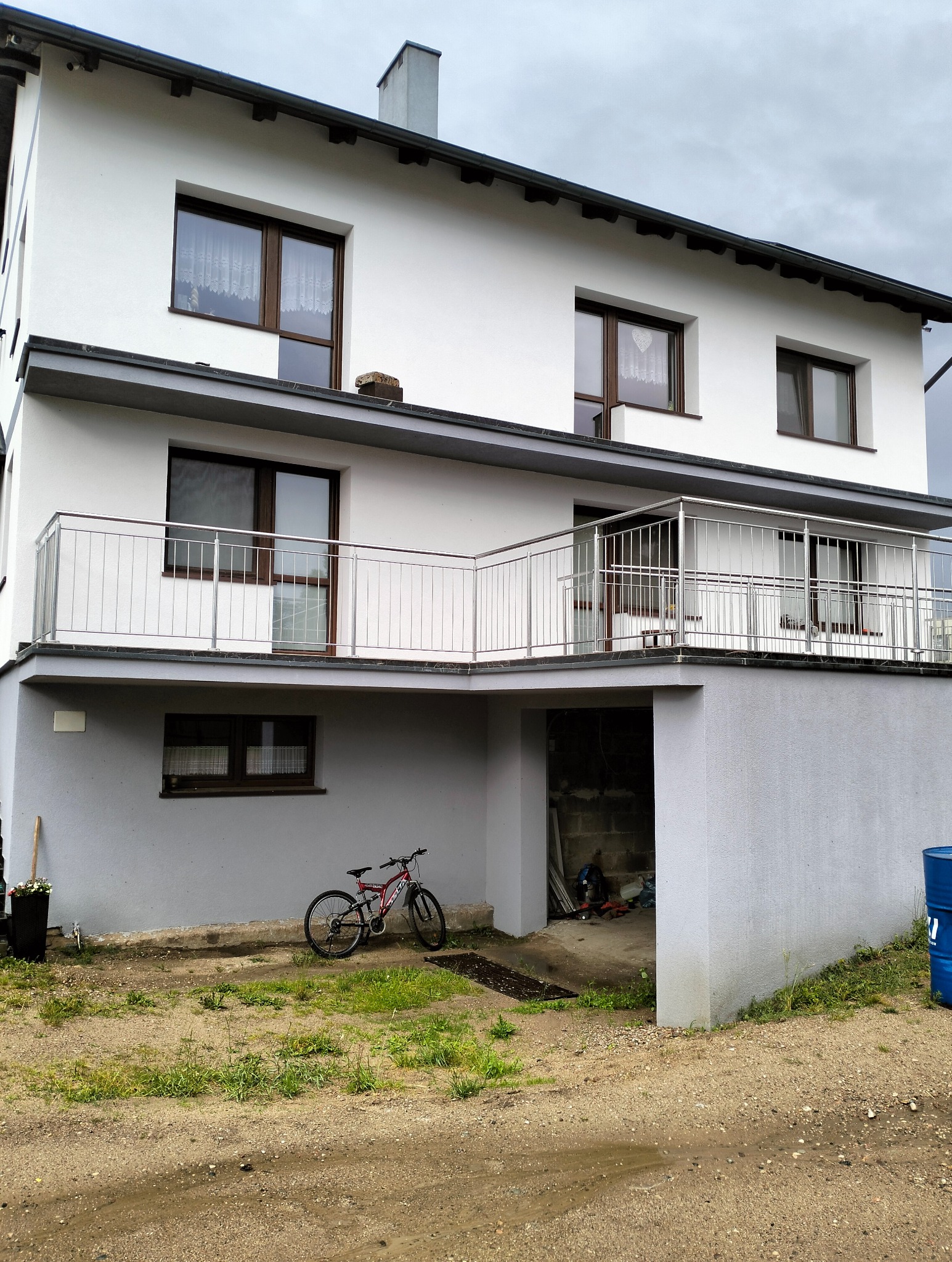 Dwupiętrowy dom z balkonem zabezpieczonym metalową balustradą, rower oparty o ścianę, widok na szarą elewację i otwarty garaż.
