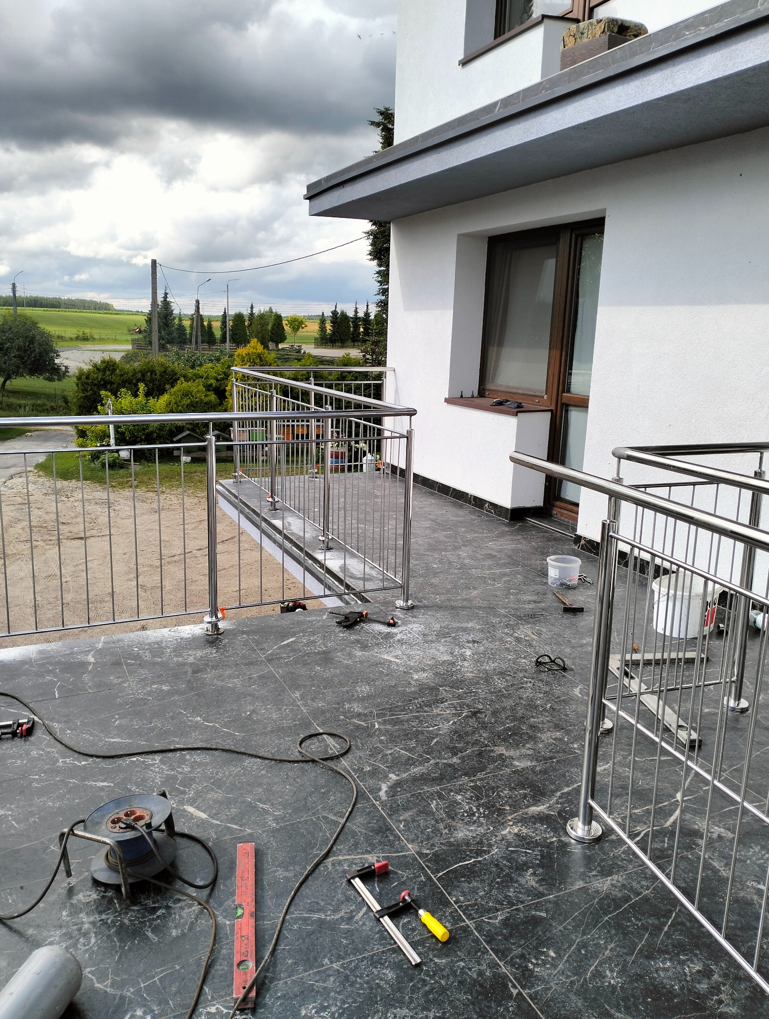 Balkon z częściowo zamontowaną balustradą ze stali nierdzewnej, widoczne narzędzia i akcesoria montażowe na marmurowej posadzce, tło stanowią drzewa i pochmurne niebo.