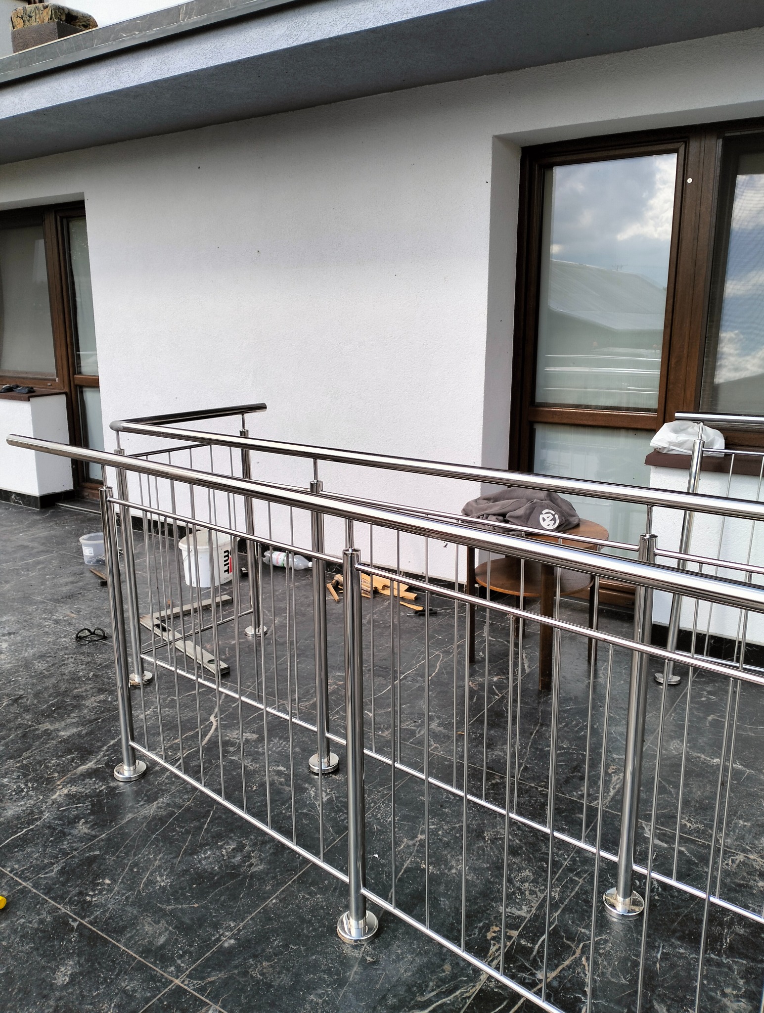 Nowoczesna balustrada ze stali nierdzewnej, zamontowana na balkonie z marmurową posadzką, widoczne narzędzia pracy, wiadro z farbą i stolik z ubraniem.