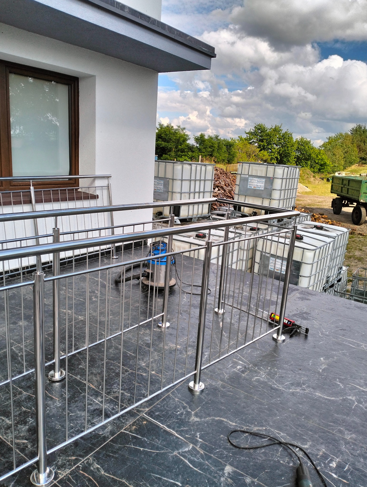 Nowoczesna balustrada ze stali nierdzewnej z cienkimi prętami pionowymi, zamontowana na tarasie wykończonym czarnym marmurem, z widocznymi narzędziami pracy i zbiornikami IBC w tle.