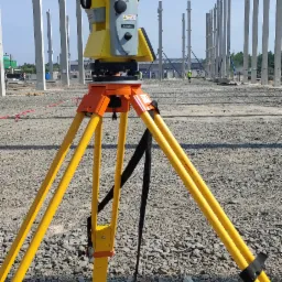 Żółto-szary teodolit na trójnogu na żwirowym terenie budowy, w tle szereg betonowych słupów konstrukcyjnych i pracownik w kamizelce odblaskowej.