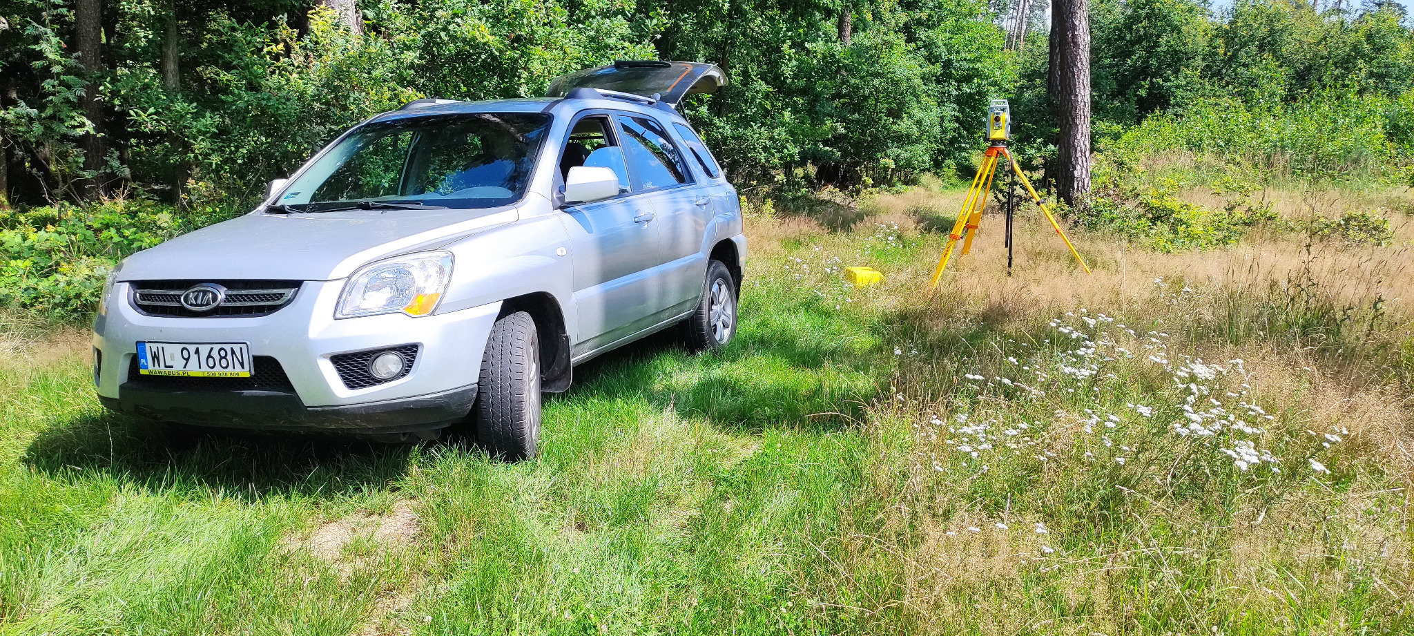 Srebrny samochód terenowy Kia Sportage z otwartym bagażnikiem zaparkowany na trawiastym terenie w pobliżu lasu; w tle żółty statyw z teodolitem.