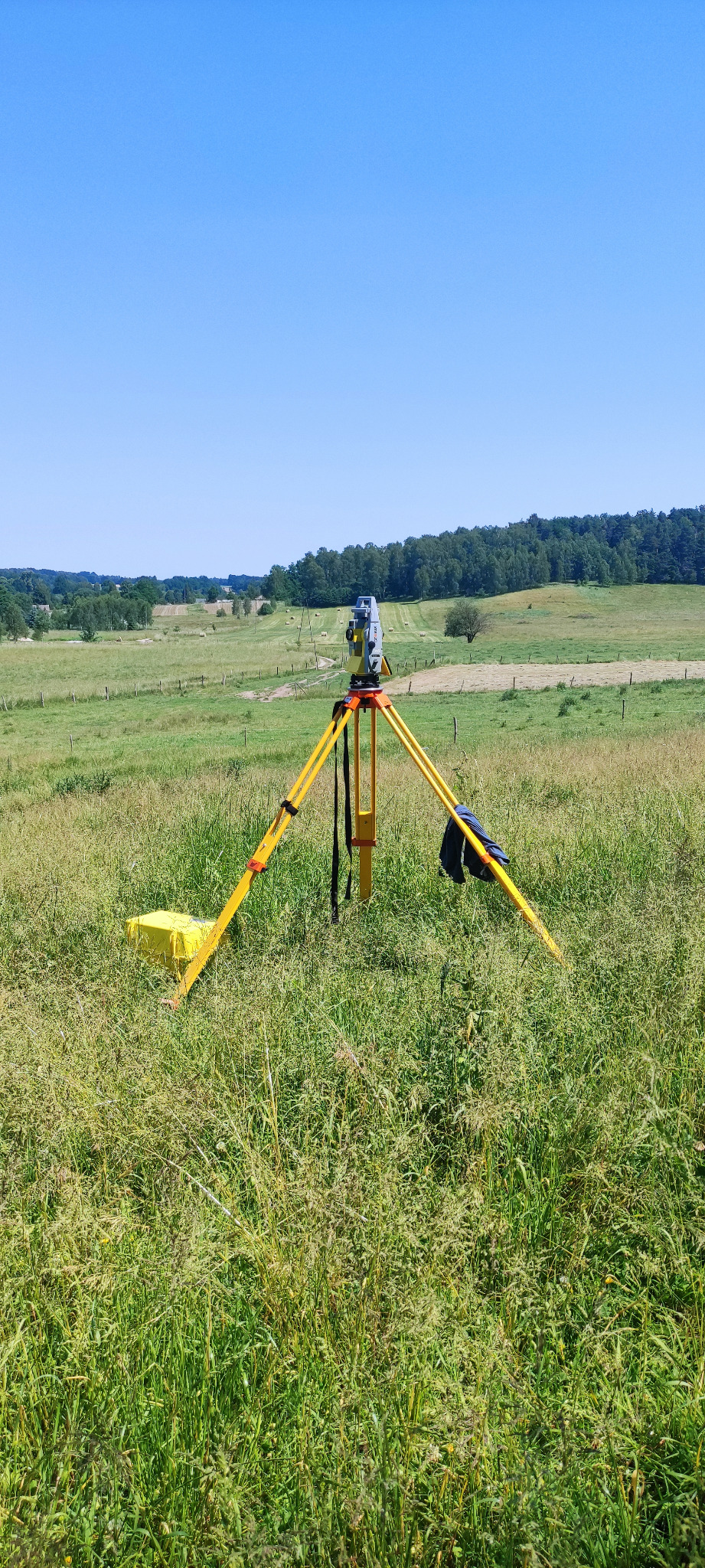 Żółty statyw z teodolitem ustawiony na zarośniętej łące z widokiem na pagórkowaty krajobraz i las w oddali.
