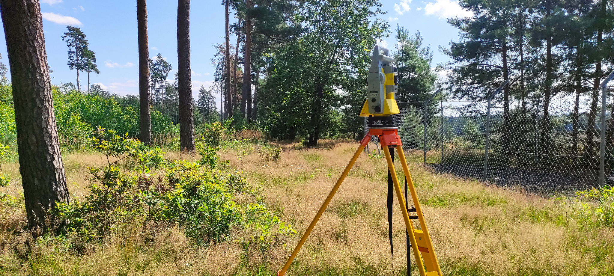 Żółto-szary teodolit ustawiony na statywie w terenie porośniętym trawą i drzewami, w tle siatka ogrodzeniowa.