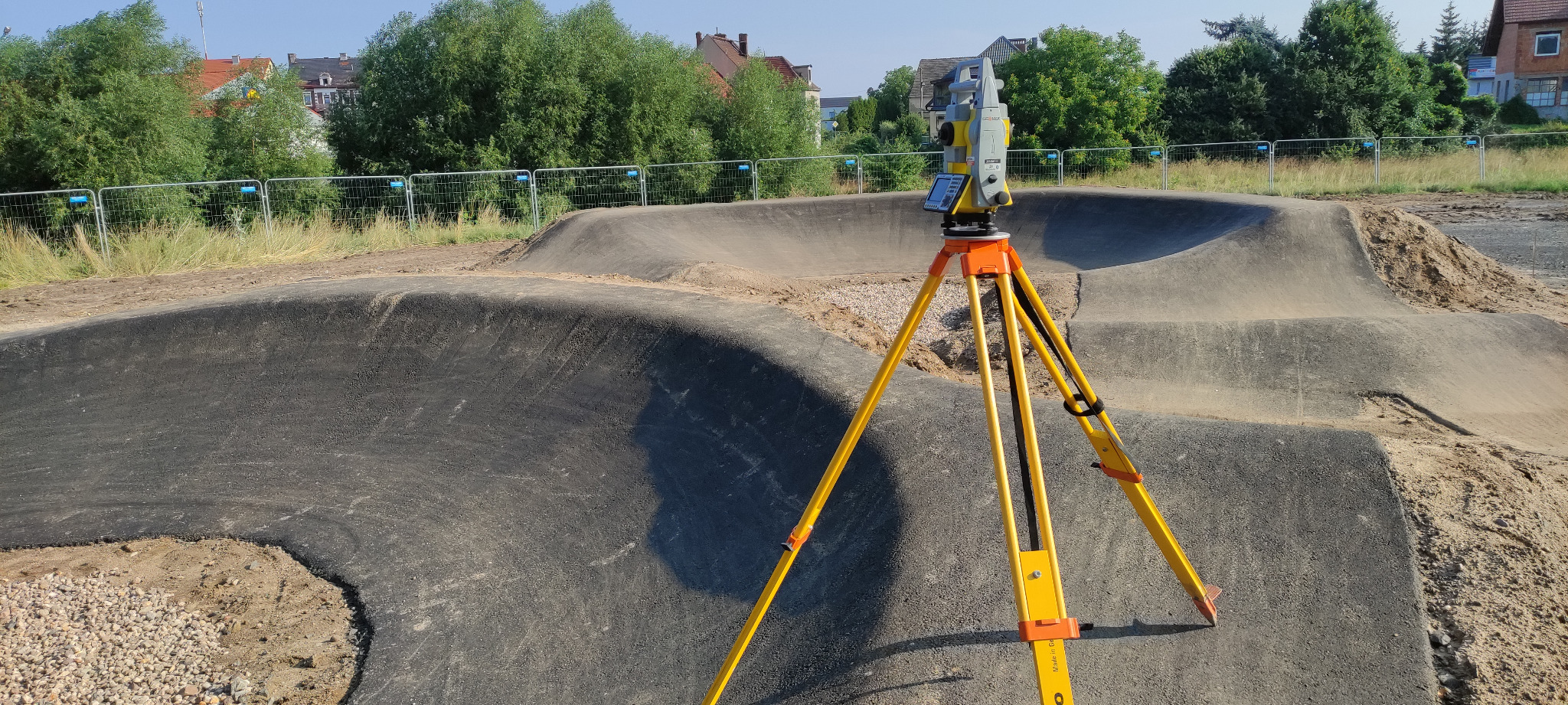 Żółty teodolit na trójnogu ustawiony na asfalcie, w tle ukształtowany teren z asfaltowymi ścieżkami, ogrodzenie z siatki i zabudowania.