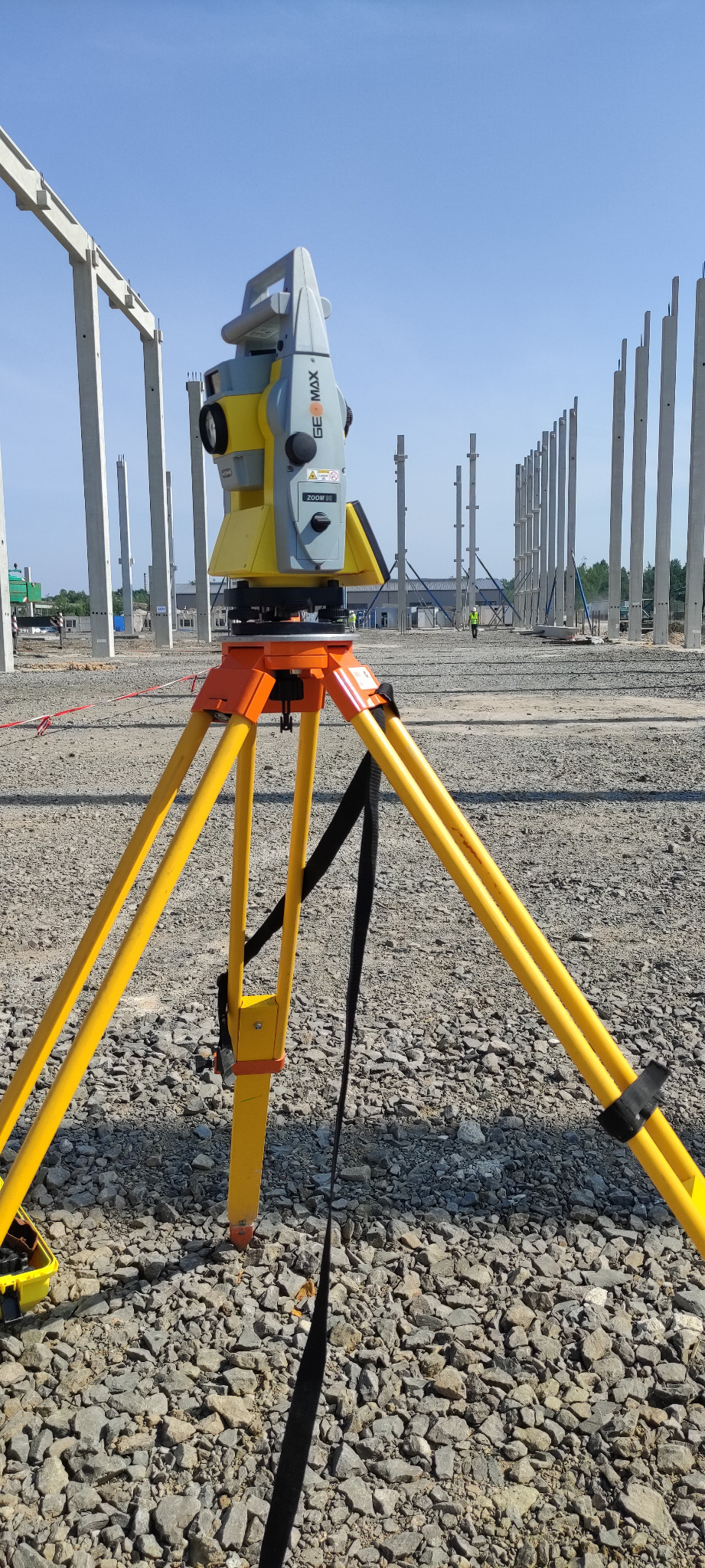 Żółto-szary teodolit na trójnogu na żwirowym terenie budowy, w tle szereg betonowych słupów konstrukcyjnych i pracownik w kamizelce odblaskowej.
