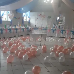 Sala bankietowa udekorowana białymi i różowymi balonami na podłodze, stoły przykryte białymi obrusami z turkusowymi i różowymi wstążkami na krzesłach, niebiesko-białe drapowania pod sufitem...