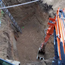 Głęboki wykop ziemny z pomarańczową koparką w trakcie pracy, widoczna drabina opuszczona do wnętrza wykopu, narzędzia i rury na dnie, ziemia o zróżnicowanej strukturze.