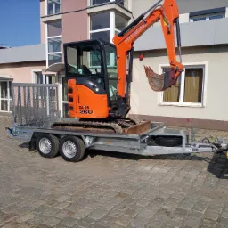 Pomarańczowa mini koparka Hitachi Zaxis 26U załadowana na szarą przyczepę z rampą, na brukowanym placu przed budynkiem.