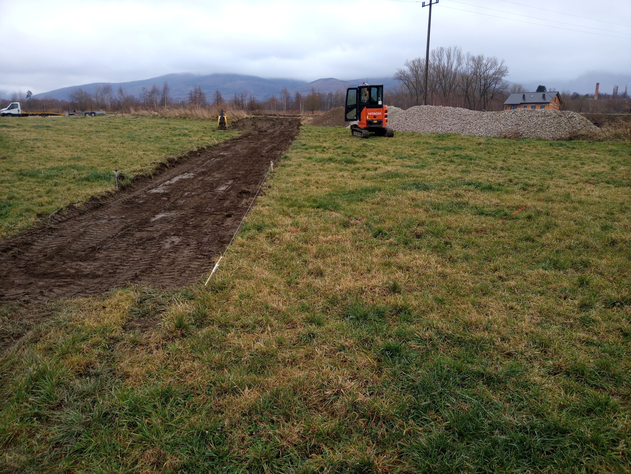 Pomarańczowa mini koparka Hitachi stoi na trawiastym polu obok wykopu z ciemną ziemią, w tle hałda żwiru i budynek, widoczne słupy wyznaczające trasę.