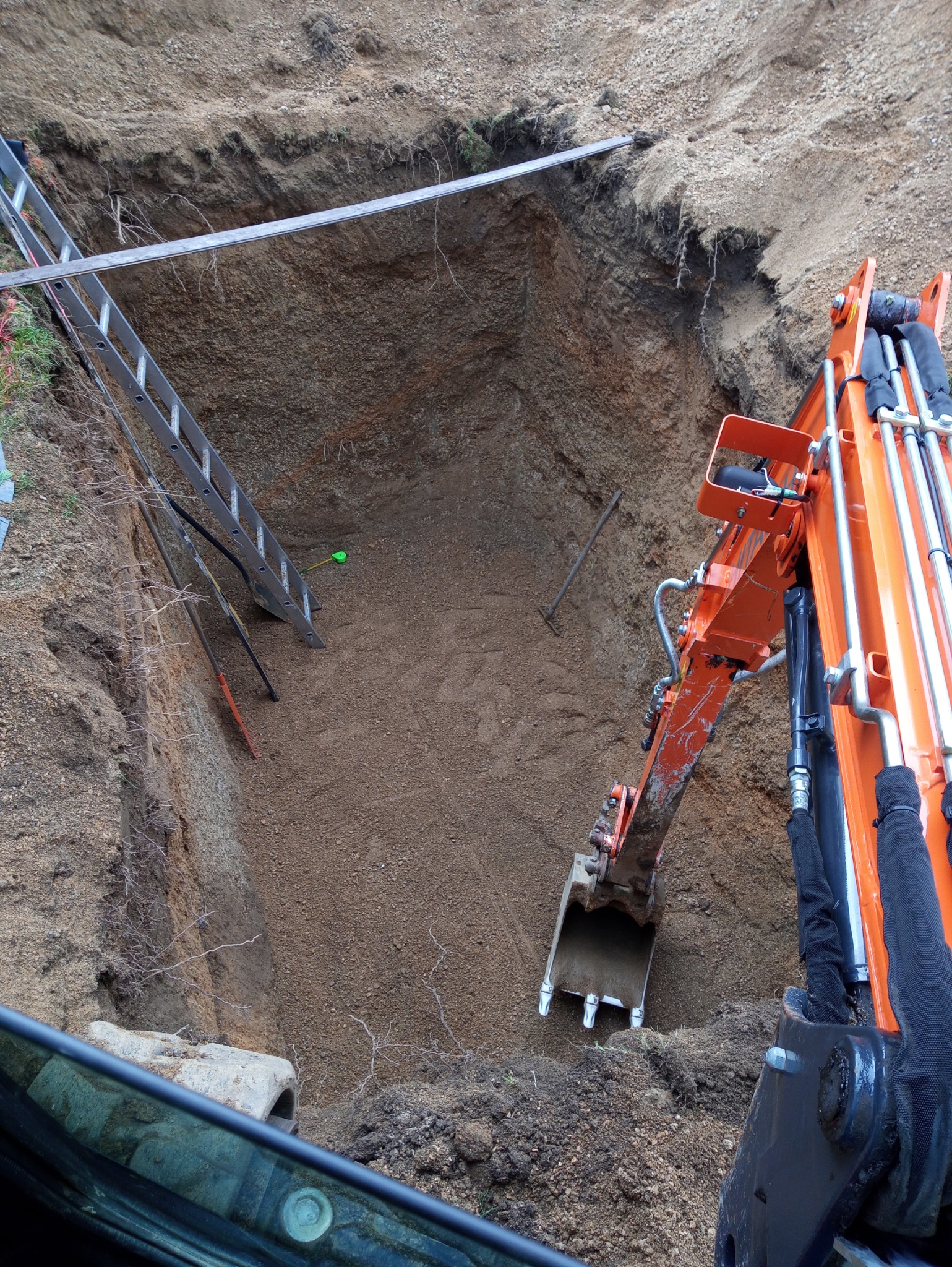 Głęboki wykop ziemny z pomarańczową koparką w trakcie pracy, widoczna drabina opuszczona do wnętrza wykopu, narzędzia i rury na dnie, ziemia o zróżnicowanej strukturze.