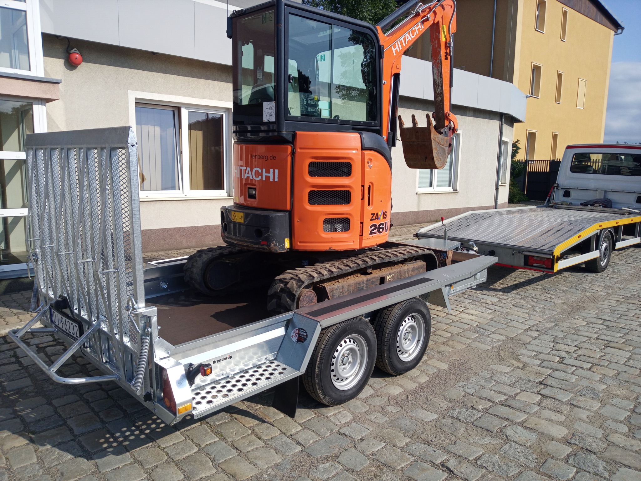 Pomarańczowa mini koparka Hitachi Zaxis 26U na srebrnej przyczepie transportowej z najazdami, zaparkowana na brukowanej nawierzchni przed budynkiem.