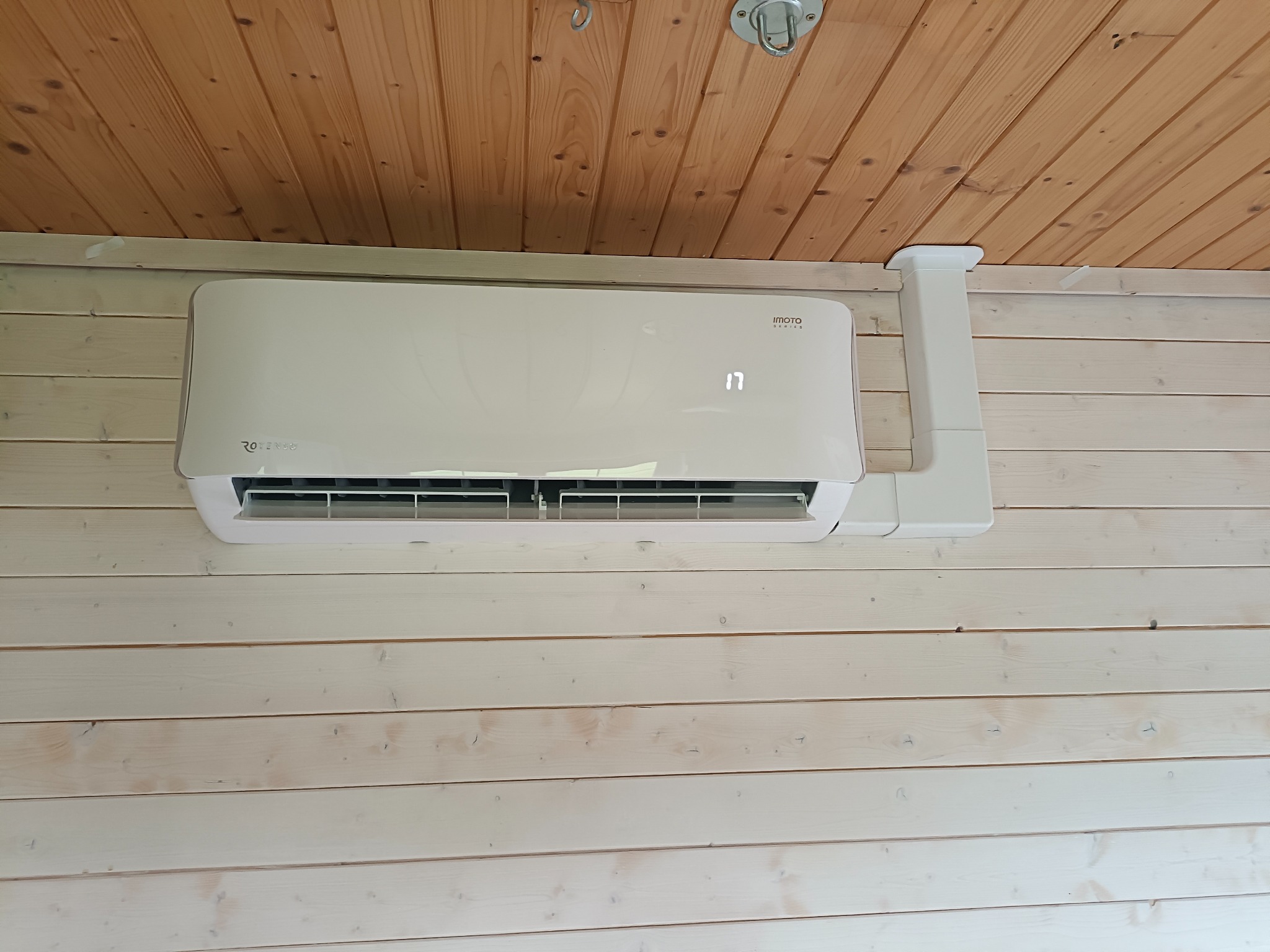 Biała jednostka klimatyzacji zamontowana na ścianie z jasnego drewna, widoczna instalacja odprowadzająca, wyświetlacz pokazuje temperaturę 17 stopni.