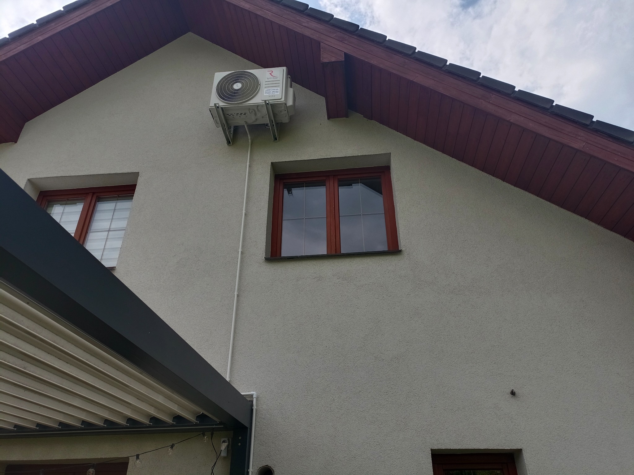 Biała jednostka zewnętrzna klimatyzacji zamontowana na elewacji domu z beżowym tynkiem obok okna z brązową ramą, widoczna biała rurka instalacyjna.