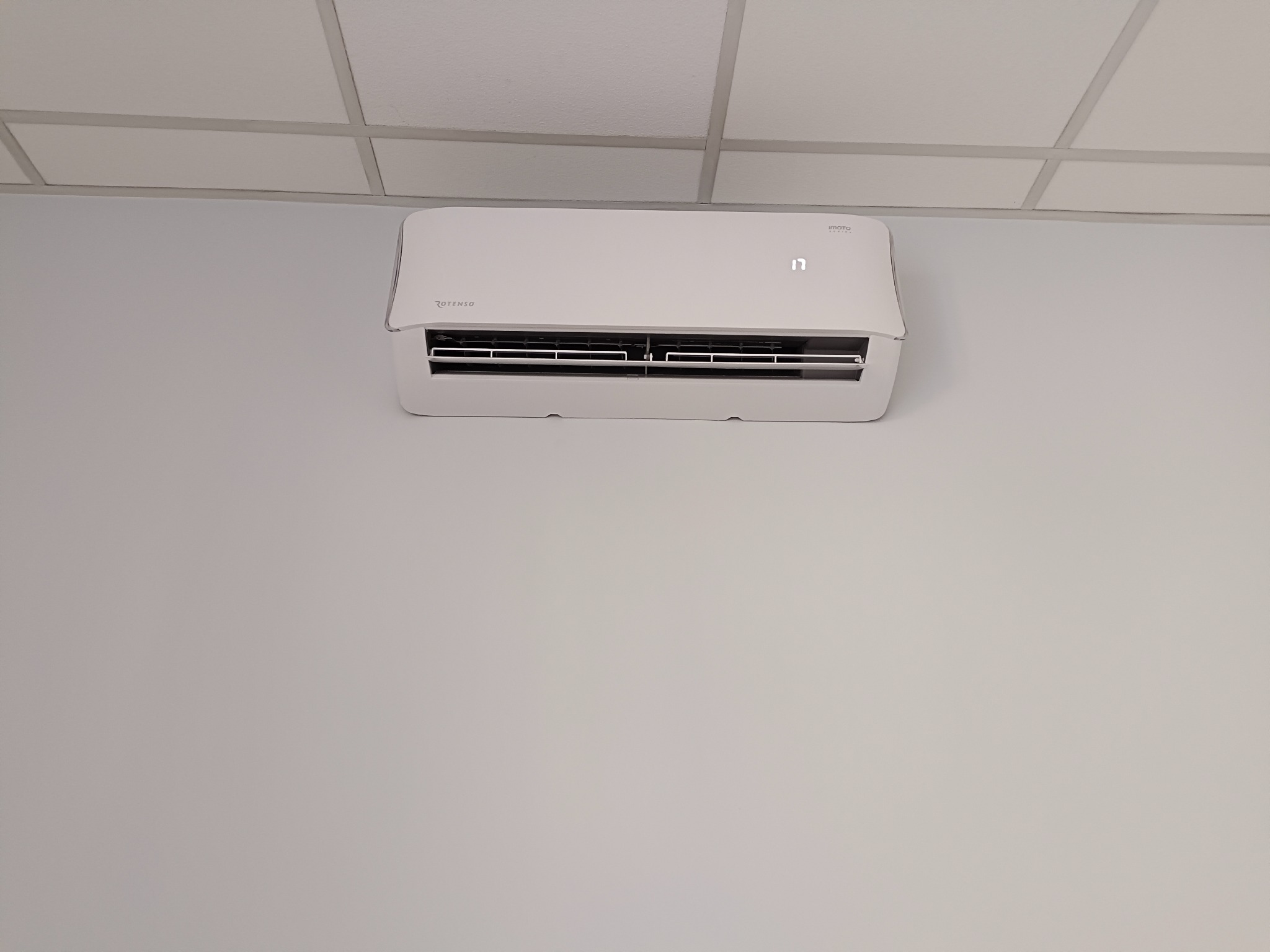 Biała jednostka klimatyzacji Rotenso Imoto, zamontowana na ścianie w pomieszczeniu z kasetonowym sufitem, wyświetlająca temperaturę 17 stopni.