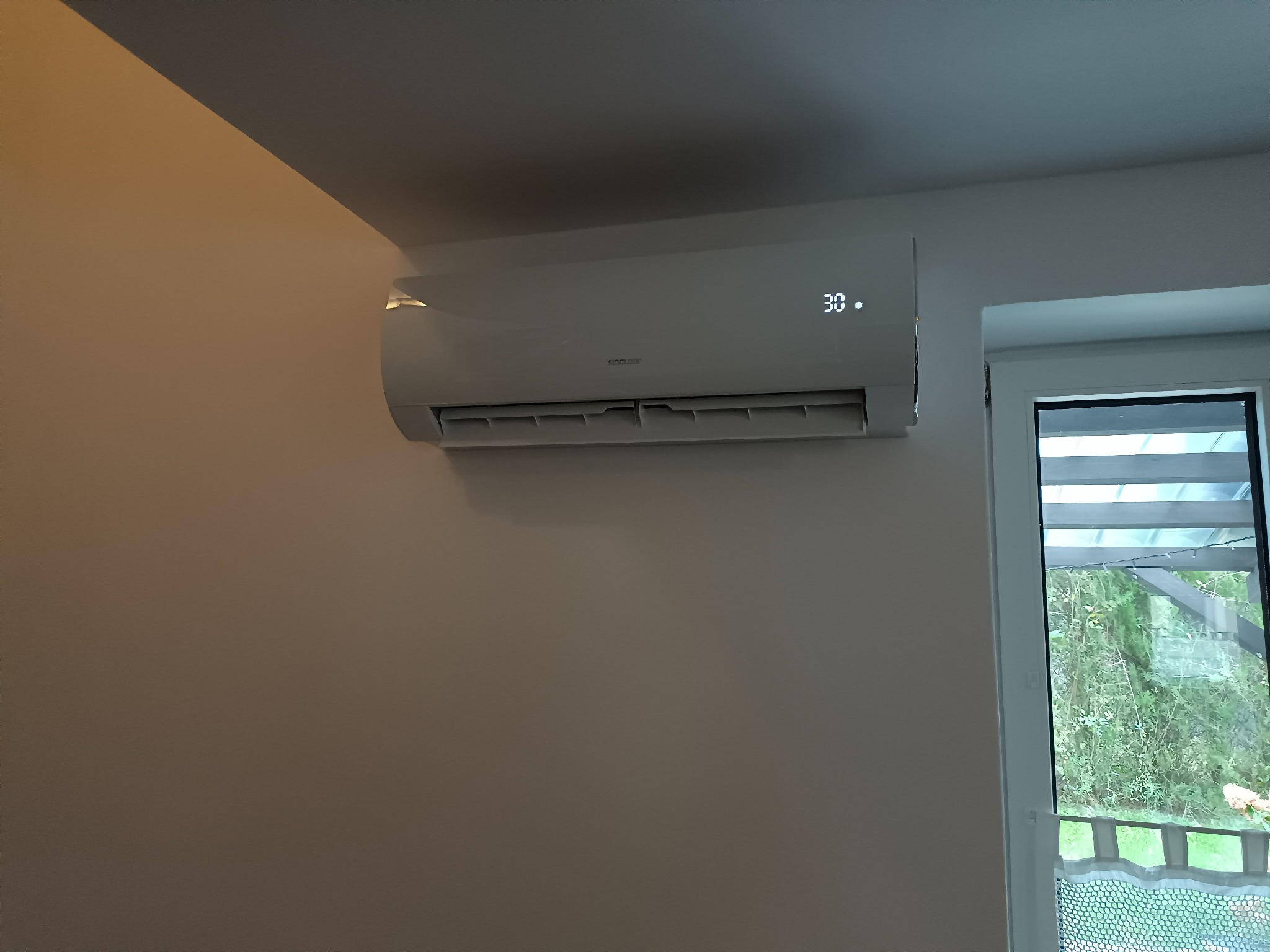 Biały klimatyzator ścienny marki Sinclair z wyświetlaną temperaturą 30 stopni Celsjusza, zamontowany w jasnym pomieszczeniu obok okna z widokiem na zieleń.