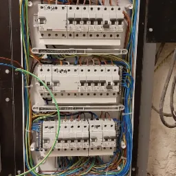 Otwarta skrzynka rozdzielcza z zamontowanymi bezpiecznikami i uporządkowanymi przewodami w różnych kolorach, widoczne ślady wiercenia i drobne zanieczyszczenia na podłożu.