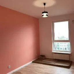 Pusty pokój po odświeżeniu: jedna ściana pomalowana na łososiowy kolor, reszta na jasny róż, drewnopodobna podłoga, czarna lampa sufitowa, okno z widokiem na budynki, kartony na podłodze.
