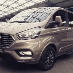 Beżowy Ford Transit Custom z czarnymi felgami, sfotografowany pod zadaszeniem z metalową konstrukcją, z widocznym fragmentem przedniego reflektora i logo Forda.