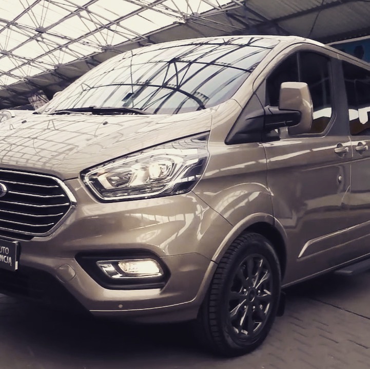 Beżowy Ford Transit Custom z czarnymi felgami, sfotografowany pod zadaszeniem z metalową konstrukcją, z widocznym fragmentem przedniego reflektora i logo Forda.