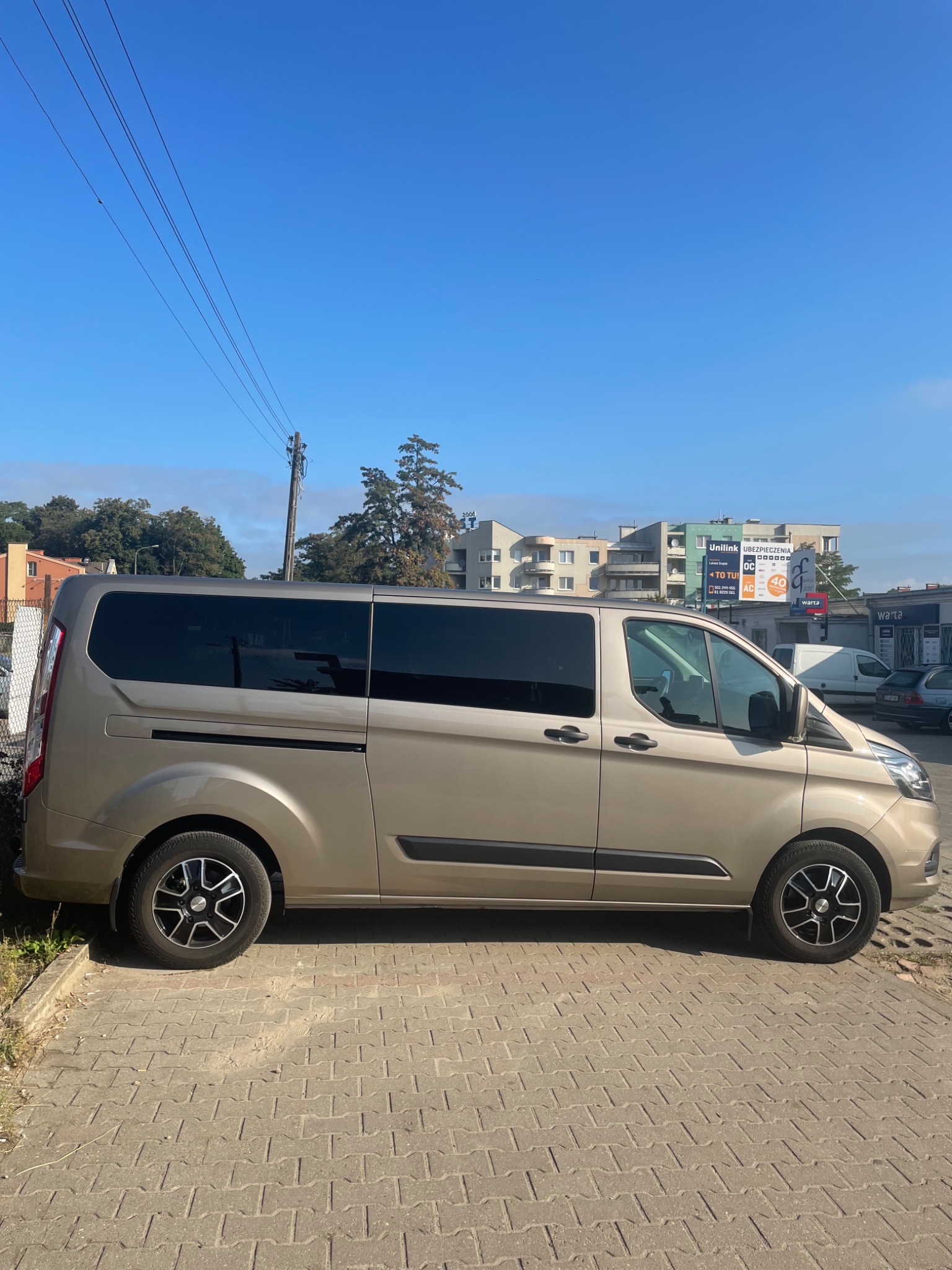 Złoty bus z przyciemnianymi szybami zaparkowany na brukowanym parkingu w słoneczny dzień, budynki i reklamy w tle.