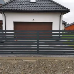 Nowoczesna, pozioma brama przesuwna w kolorze grafitowym, zamontowana na posesji z domem o białej elewacji i brązową bramą garażową, widok z zewnątrz na żwirową nawierzchnię.