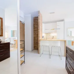 Jasne wnętrze mieszkania z widokiem na kuchnię i jadalnię, drewniana komoda z lustrem w ramie, białe płytki podłogowe.