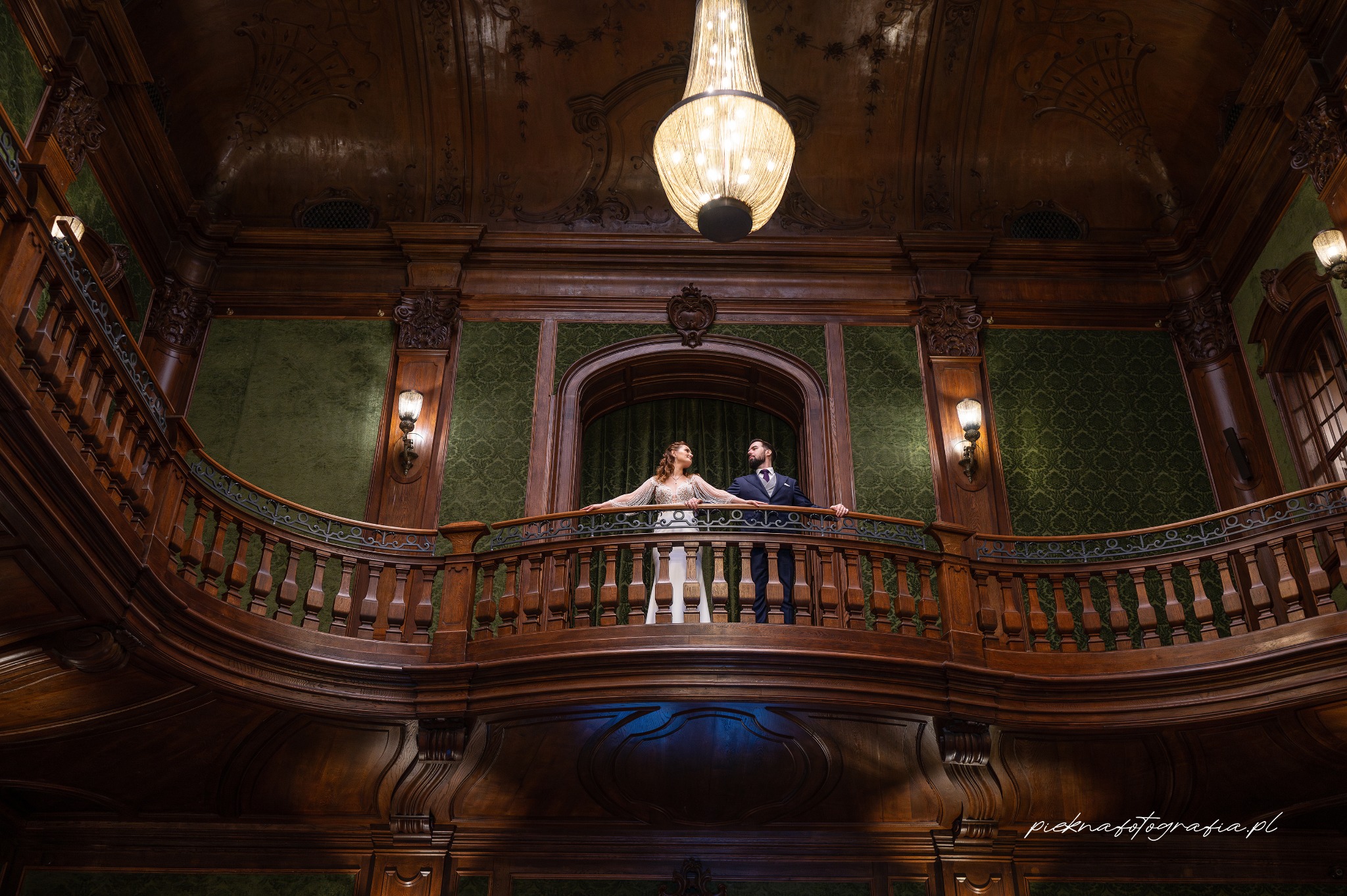 Para młoda na drewnianym balkonie w bogato zdobionym wnętrzu z żyrandolem, on w granatowym garniturze, ona w sukni ślubnej z długimi rękawami, oboje patrzą na siebie.