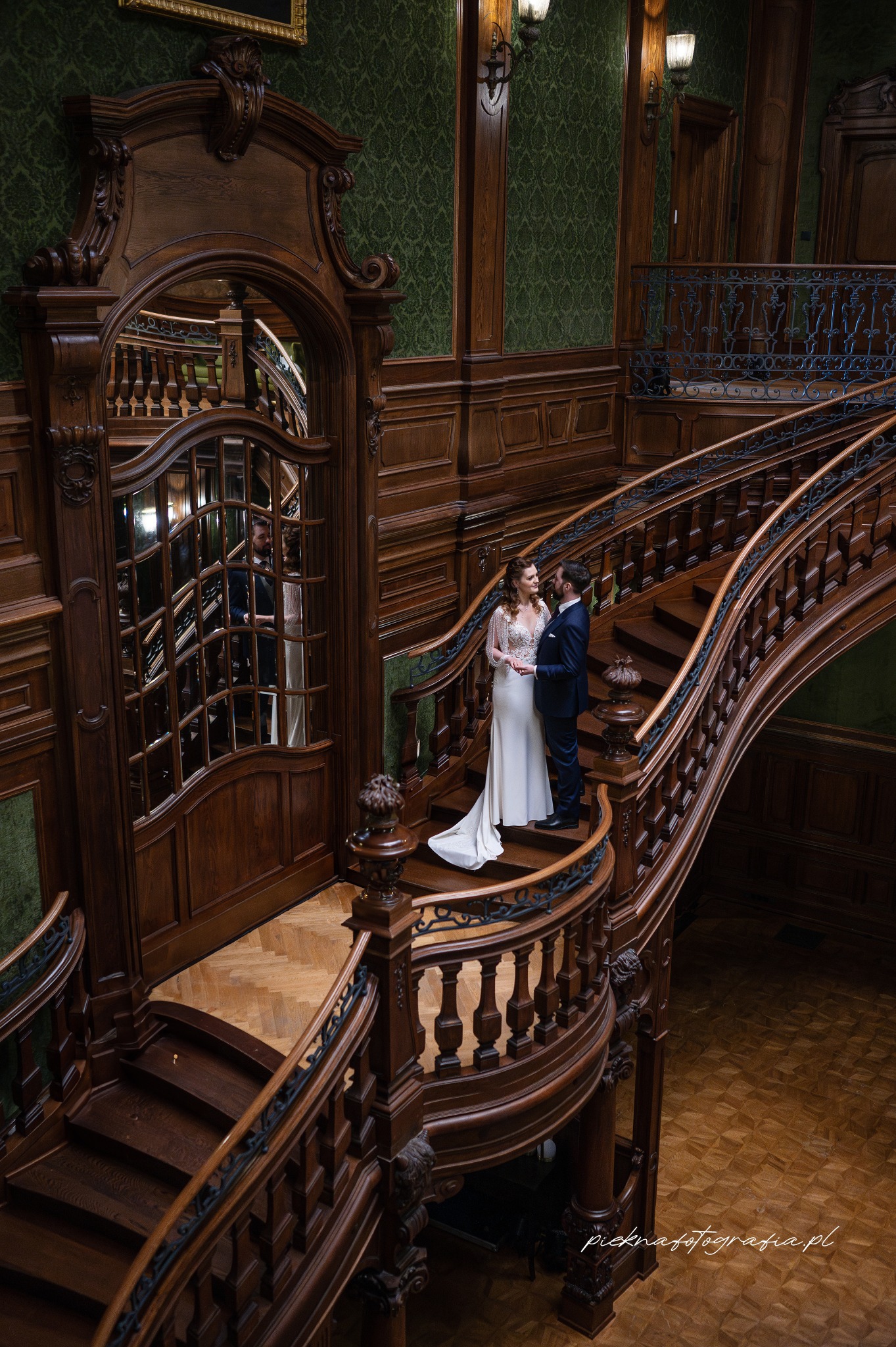 Para młoda na monumentalnych, drewnianych schodach w eleganckim wnętrzu z zielonymi ścianami i ozdobnymi detalami.