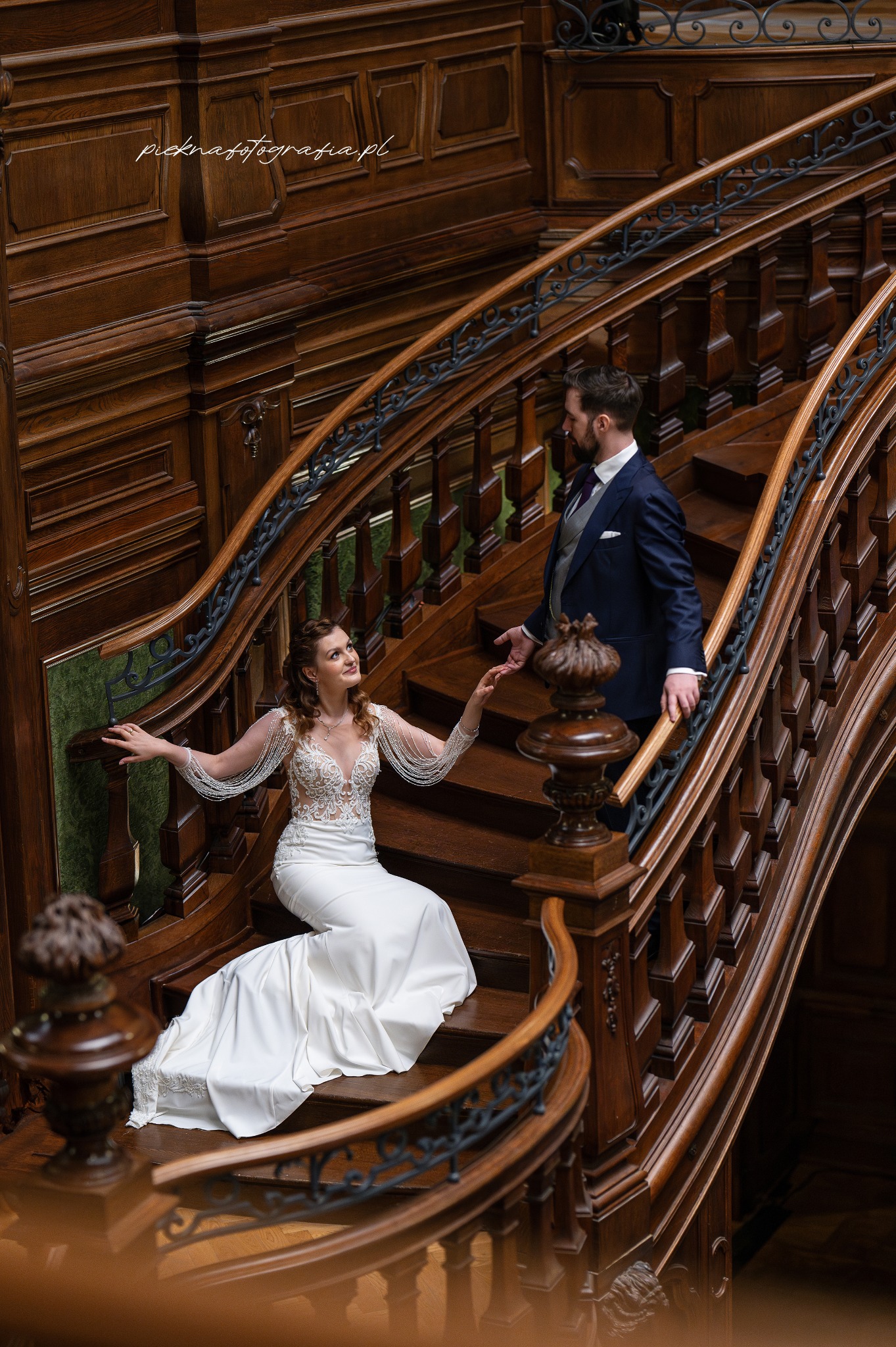 Panna młoda w białej sukni z koronkową górą i ozdobnymi rękawami siedzi na drewnianych schodach, spoglądając na pana młodego w granatowym garniturze. Schody z ciemnego drewna z ozdobnymi...