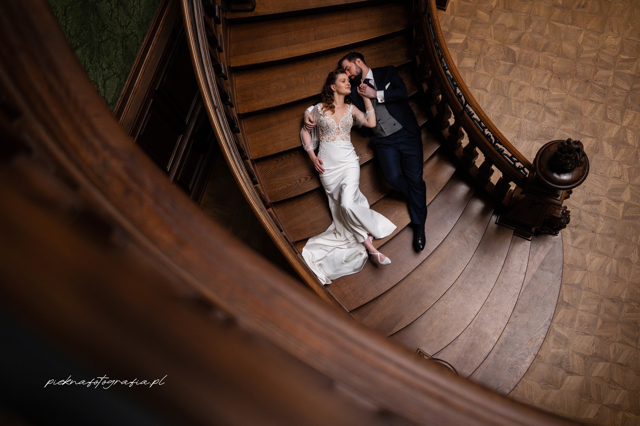 Para młoda leżąca na drewnianych schodach z ozdobną balustradą, panna młoda w białej sukni z koronkową górą, pan młody w granatowym garniturze z szarą kamizelką, widok z góry.