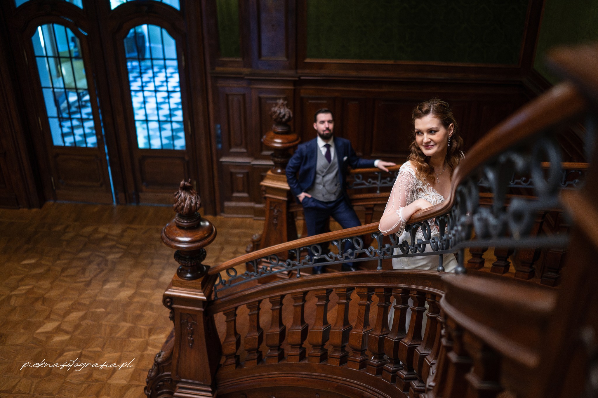 Para młoda na drewnianych schodach w zabytkowym wnętrzu: panna młoda w koronkowej sukni z koralikami, pan młody w granatowym garniturze i szarej kamizelce.