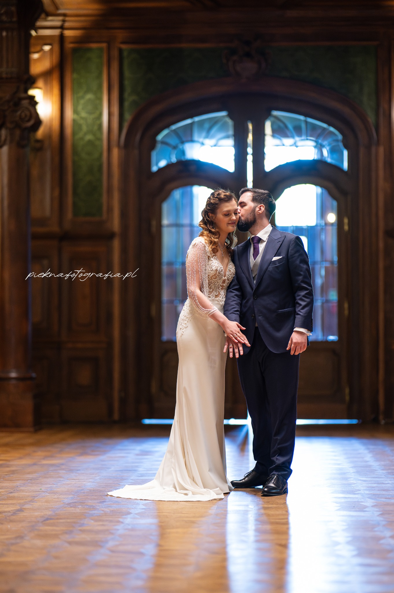 Panna młoda w sukni z koronkową górą i pan młody w granatowym garniturze, całujący się na tle bogato zdobionych drewnianych drzwi w przestronnym wnętrzu.