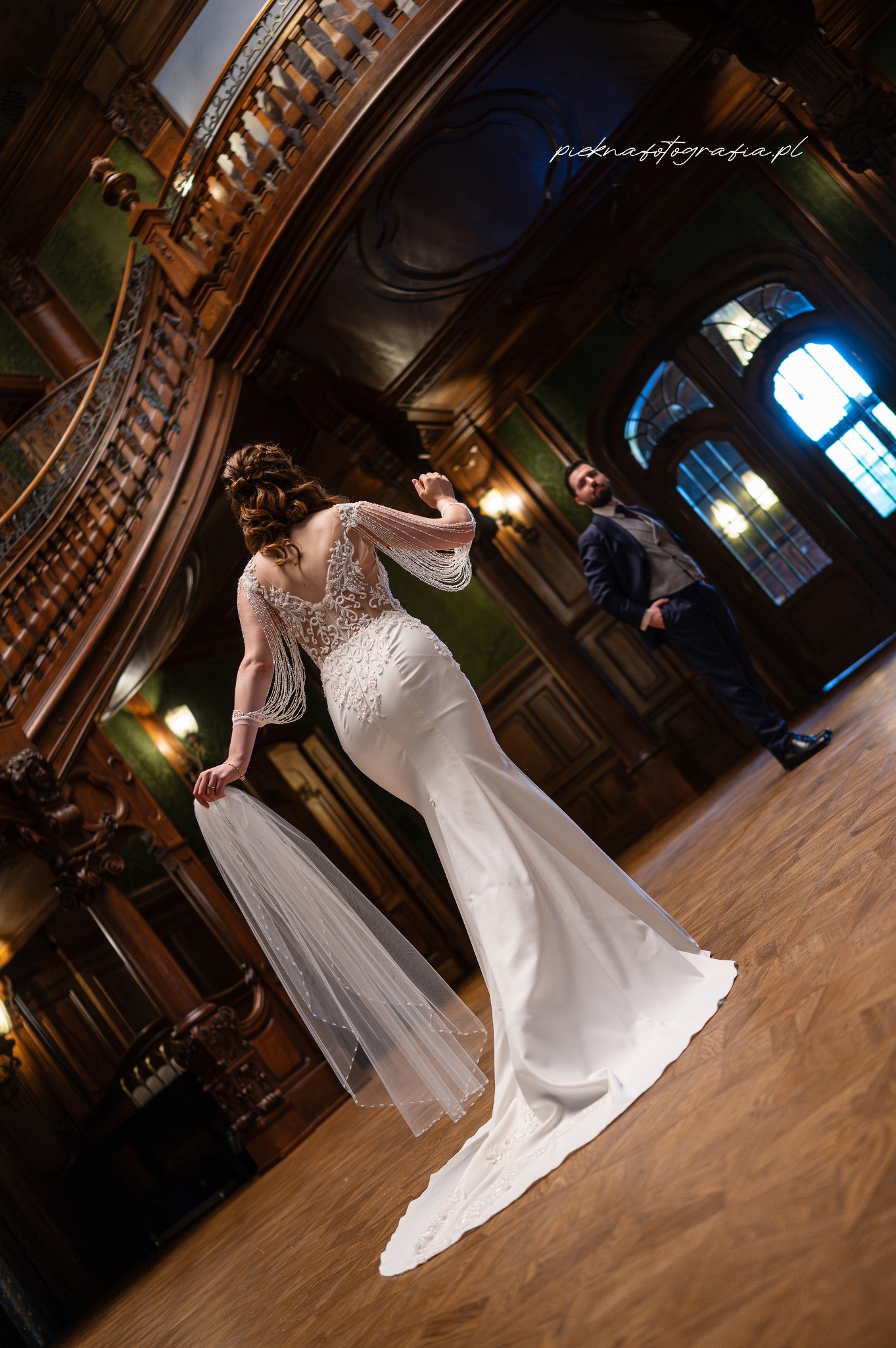 Panna młoda w koronkowej sukni z koralikami, trzymająca welon, pozuje na tle drewnianych schodów i drzwi; w tle widoczny pan młody w garniturze.