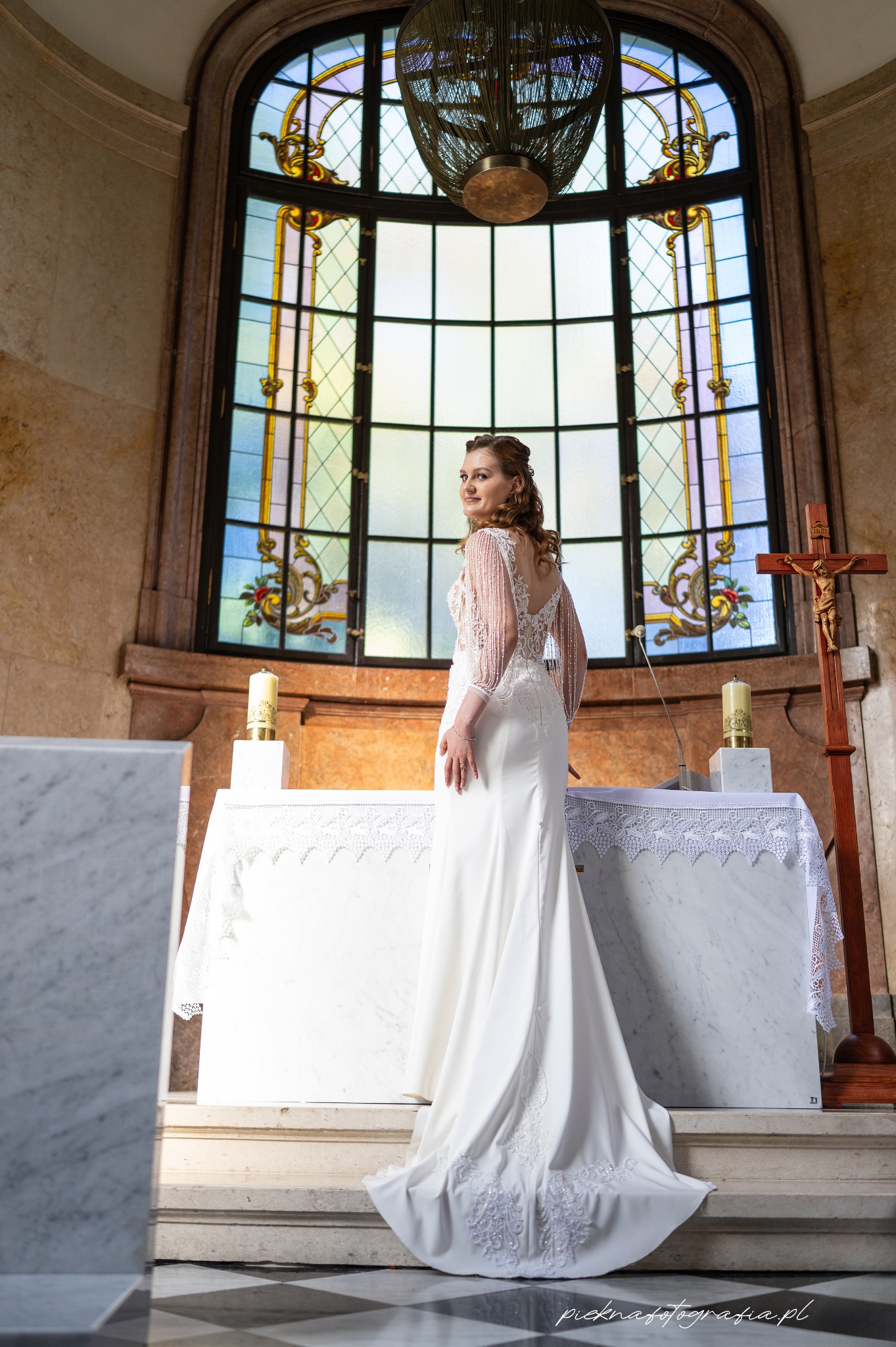 Panna młoda w białej sukni z koronkowymi rękawami, stojąca przed ołtarzem w kościele z witrażowym oknem w tle, odwrócona przez ramię.